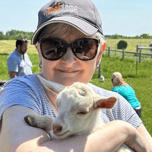 A woman wearing sunglasses and a ball cap smiles while holding a small white goat in her arms on a sunny day outdoors.