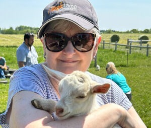 A woman wearing sunglasses and a ball cap smiles while holding a small white goat in her arms on a sunny day outdoors.