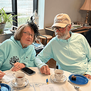 AAn older couple sits at a round table with tea cups, treats, and voting sheets in front of them during the garden photo contest. The woman wears an aqua hoodie and black fascinator and the man wears glasses and a Beyond Housing cap.