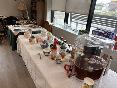 Table covered with a white cloth holding teapots, cups, treats, and a large drink dispenser, set up for a community tea gathering beside a bright window.