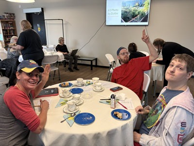 Three residents sit at a round table with teacups and snacks, smiling and waving toward the camera during a community event.