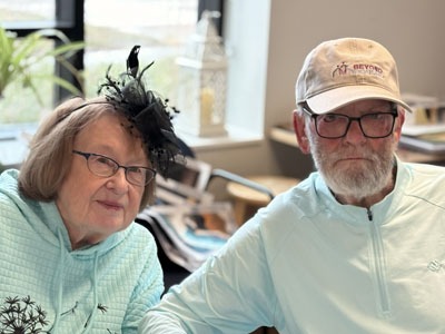 An older woman in a light blue hoodie and black fascinator sits beside an older man wearing glasses and a Beyond Housing cap.