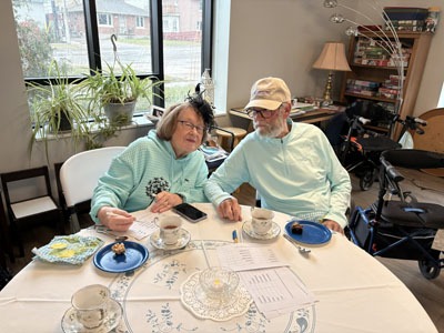 An older couple sits at a round table with tea cups, treats, and voting sheets in front of them during the garden photo contest.
