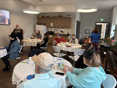 Residents sit at round tables enjoying tea and treats while watching a slideshow in a bright community room.
