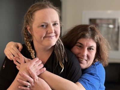 Two women smile warmly as one wraps her arms around the other in a friendly hug.