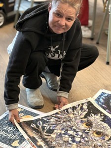 Woman crouches on the floor holding up sections of completed beadwork a detailed owl design.