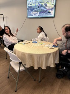 Three residents sit around a round table with teacups while a garden photo slideshow plays on the screen behind them.