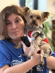 Woman in a blue shirt holds a small Yorkshire terrier wearing a red harness, looking toward the camera.