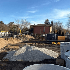 A sunny construction site with piles of dirt and gravel, concrete pipes, and machinery scattered across the area, with houses and leafless trees in the background.