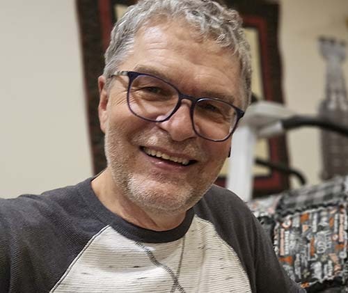 A smiling man with short grey hair and glasses takes a relaxed selfie indoors, with patterned fabric and exercise equipment in the background.
