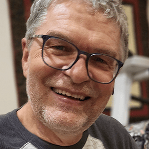 A smiling man with short grey hair and glasses takes a relaxed selfie indoors, with patterned fabric and exercise equipment in the background.