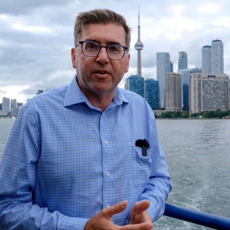A man in a blue checkered shirt stands by a waterfront railing with the Toronto skyline and CN Tower behind him under cloudy skies.