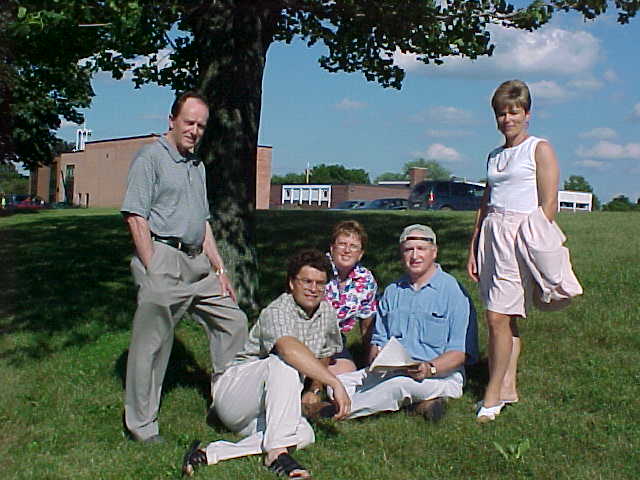 Five adults pose together outdoors on a sunny day, with two seated on the grass and three standing under a large tree, with a school and church in the background.