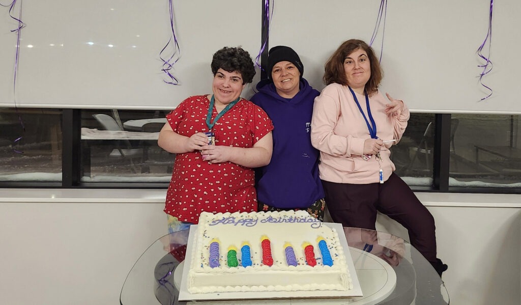 Three women stand behind a table with a birthday cake on it.