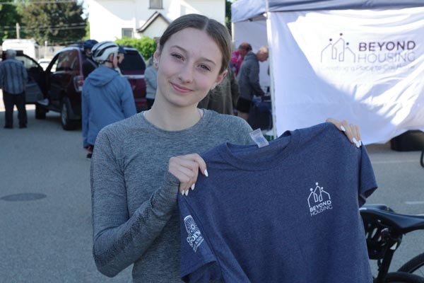 Young woman holds a Beyond Housing T-shirt at an Out-Spok’n Bike-a-Thon event while participants gather near a Beyond Housing tent in the background.