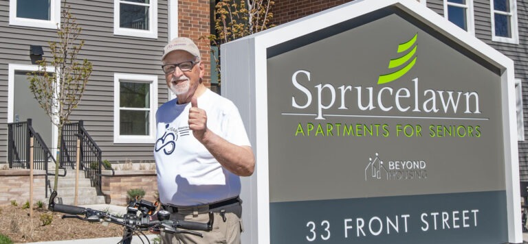 An older man on a bicycle stands next to a sign reading Sprucelawn Apartments for Seniors