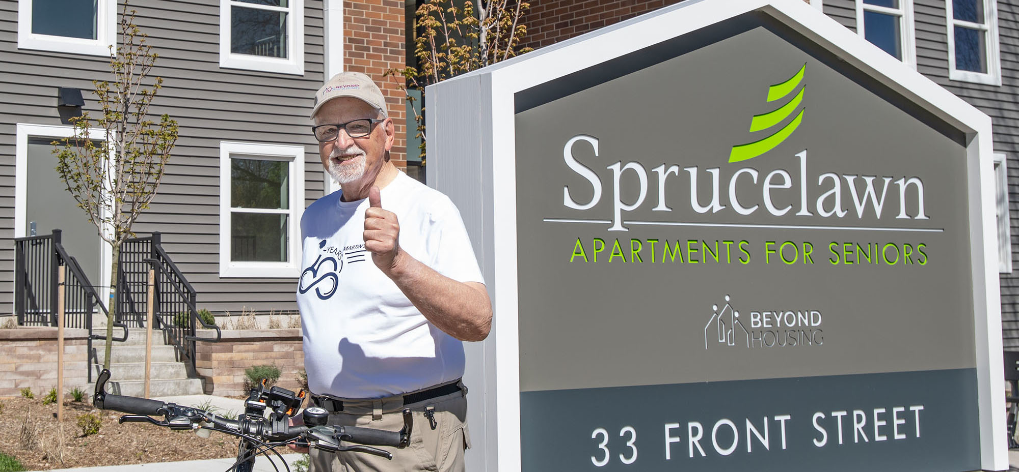 An older man on a bicycle stands next to a sign reading Sprucelawn Apartments for Seniors