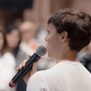 Woman speaking into a handheld microphone while addressing a small group in an indoor setting.