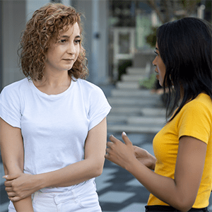 Two women standing outdoors engaged in conversation, one listening attentively while the other gestures as she speaks.