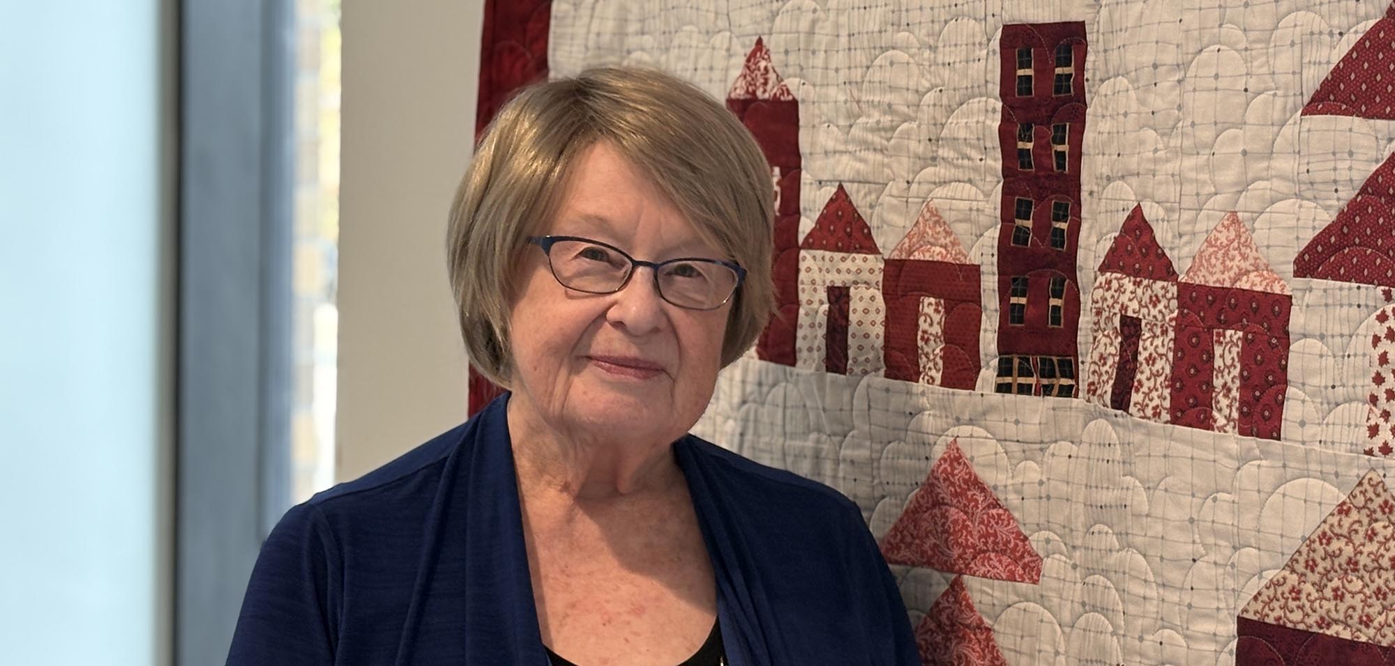 An older woman stands beside a quilt decorated with houses.