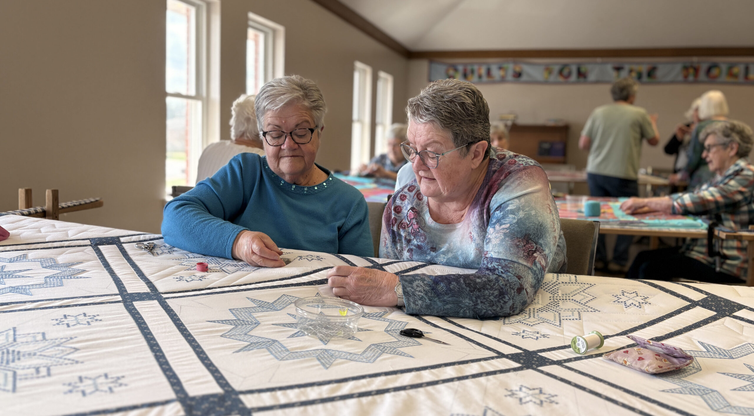 Two women concentrate on hand-stitching a quilt with blue star patterns.