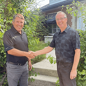 Two men standing outdoors shaking hands in front of a house, smiling at the camera.