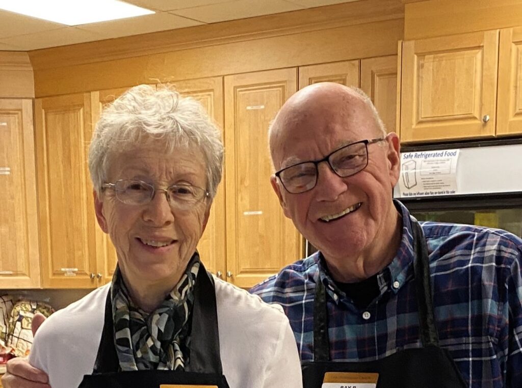 An older couple standing in front of kitchen cabinets smile at the camera.