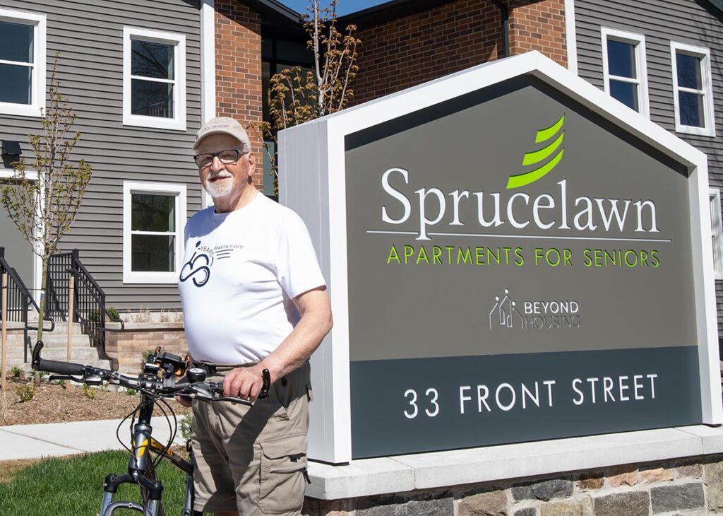 Martin Buhr stands with his bike by a sign reading Sprucelawn Apartments for Seniors