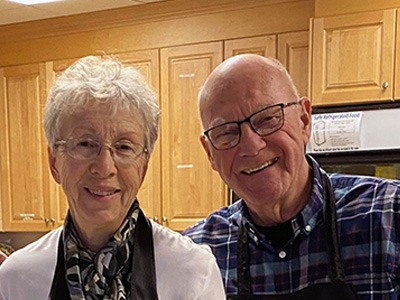 Smiling older couple standing indoors, posing together in a warmly lit kitchen setting.