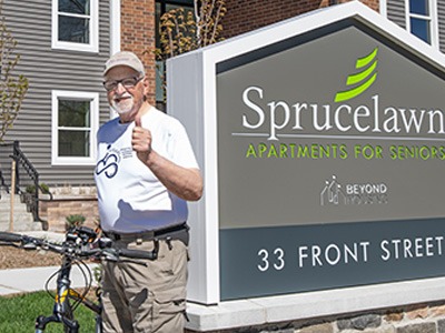 Older man standing beside a sign for Sprucelawn Apartments for Seniors, giving a thumbs-up outdoors.