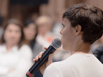Person speaking into a microphone while addressing an audience in a meeting or presentation setting.