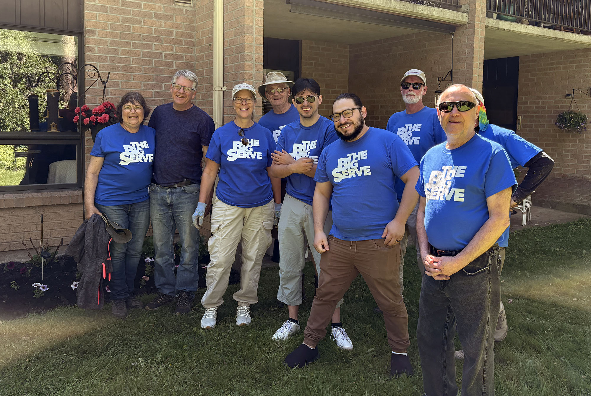 Volunteers wearing matching “The Big Serve” shirts pose together outside Sprucelawn Apartments for Seniors after completing a landscaping project.
