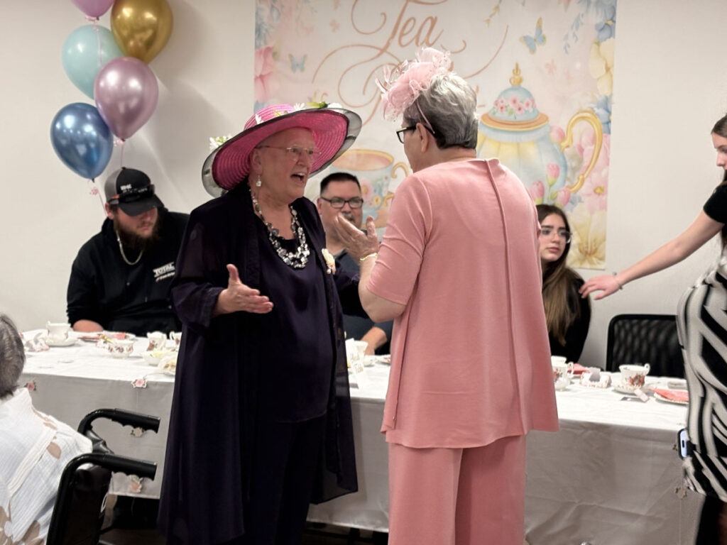Connie, wearing a large pink hat and dark outfit, chats animatedly with another tenant dressed in pink during Sprucelawn’s High Tea.