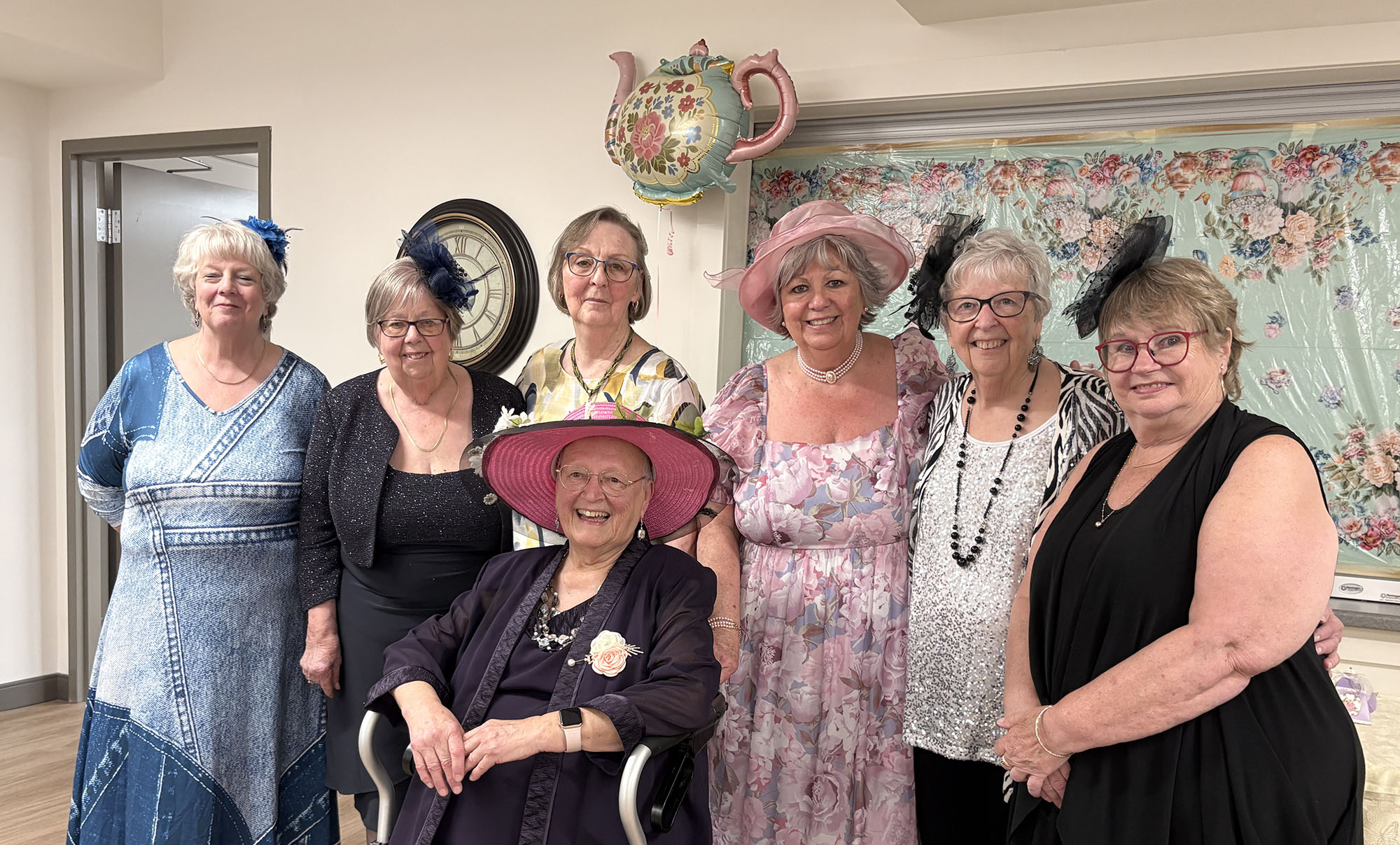 A group of guests wearing fancy hats gather below a teaoot-shaped balloon.