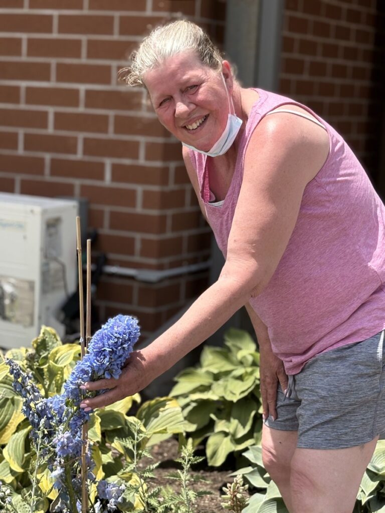 A smiling resident reaches toward blue flowers while tending a community garden outside the building.