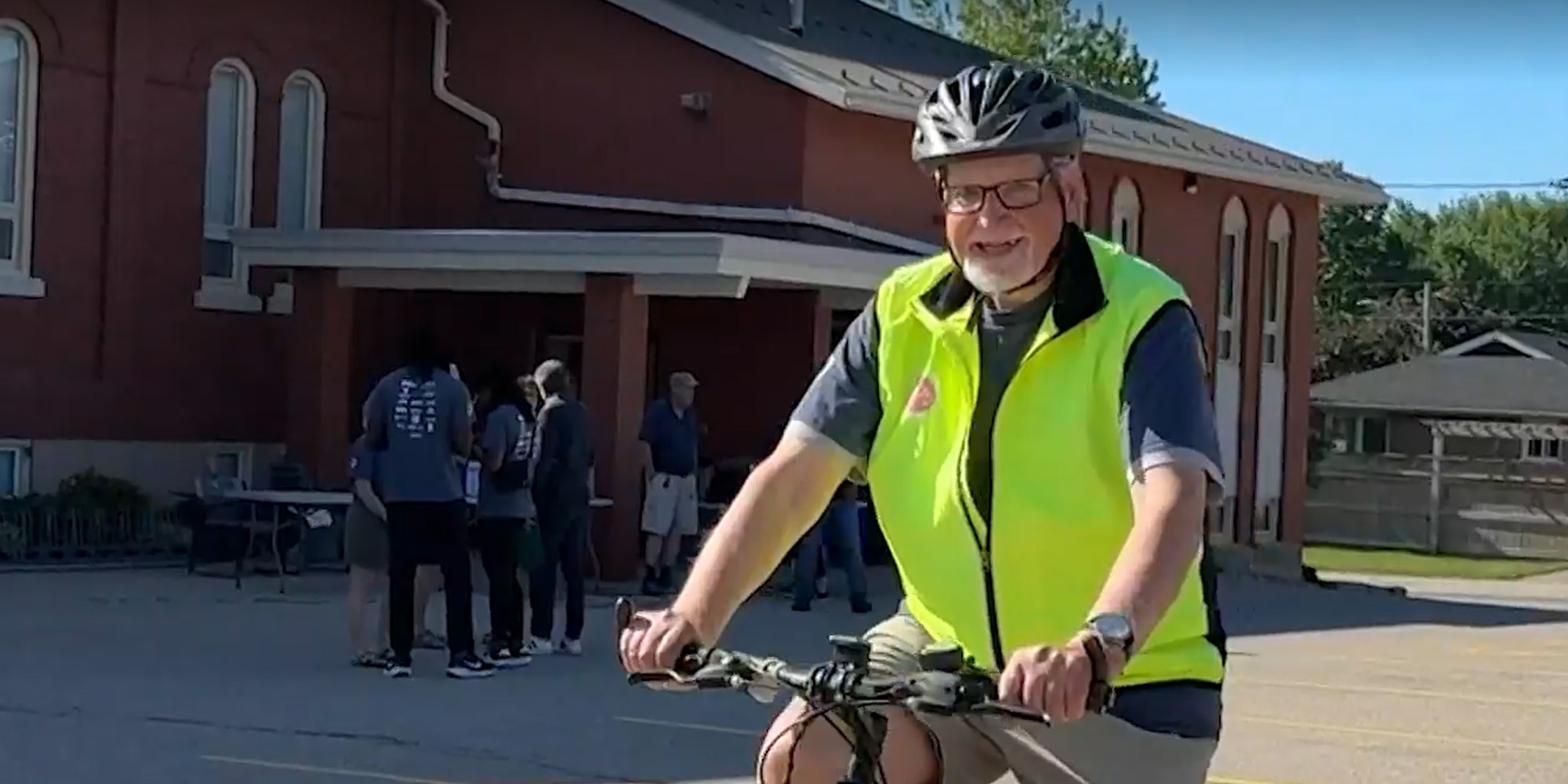 Martin, wearing a bicycle helmet and bright yellow safety vest, smiles while riding a bicycle.
