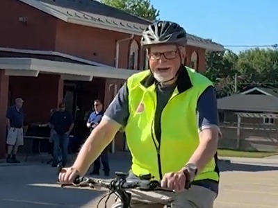 Martin, wearing a bicycle helmet and bright yellow safety vest, smiles while riding a bicycle.