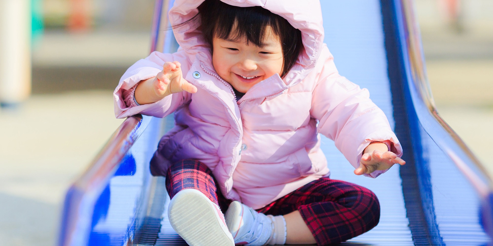Young child in a pink jacket smiles while sitting on a blue playground slide.