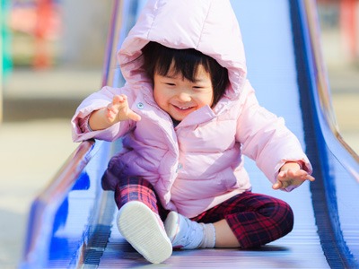 Young child in a pink jacket smiles while sitting on a blue playground slide.