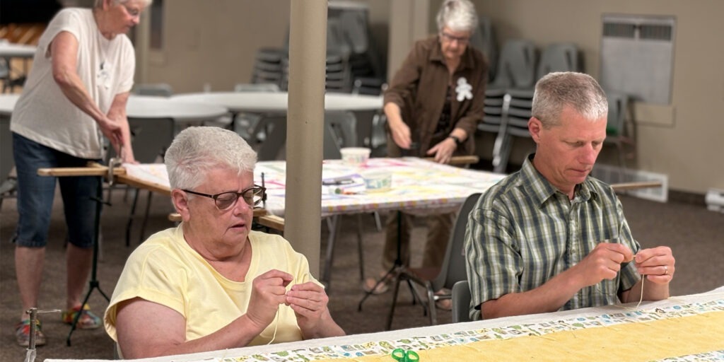 Four volunteers work together knotting colourful comforters stretched across quilting frames at Erb Street Mennonite Church.