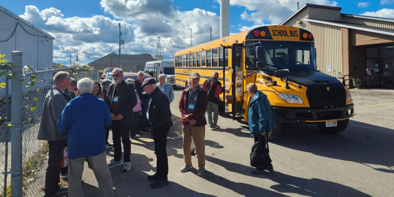 A group of Beyond Housing supporters gathers beside a yellow school bus on a sunny day during the 25th anniversary bus tour.