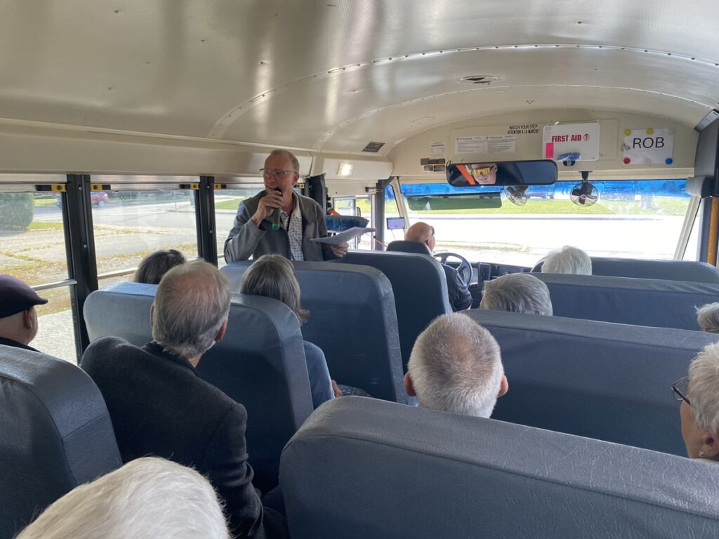 Dan speaks into a microphone at the front of a school bus as Beyond Housing bus tour guests listen.